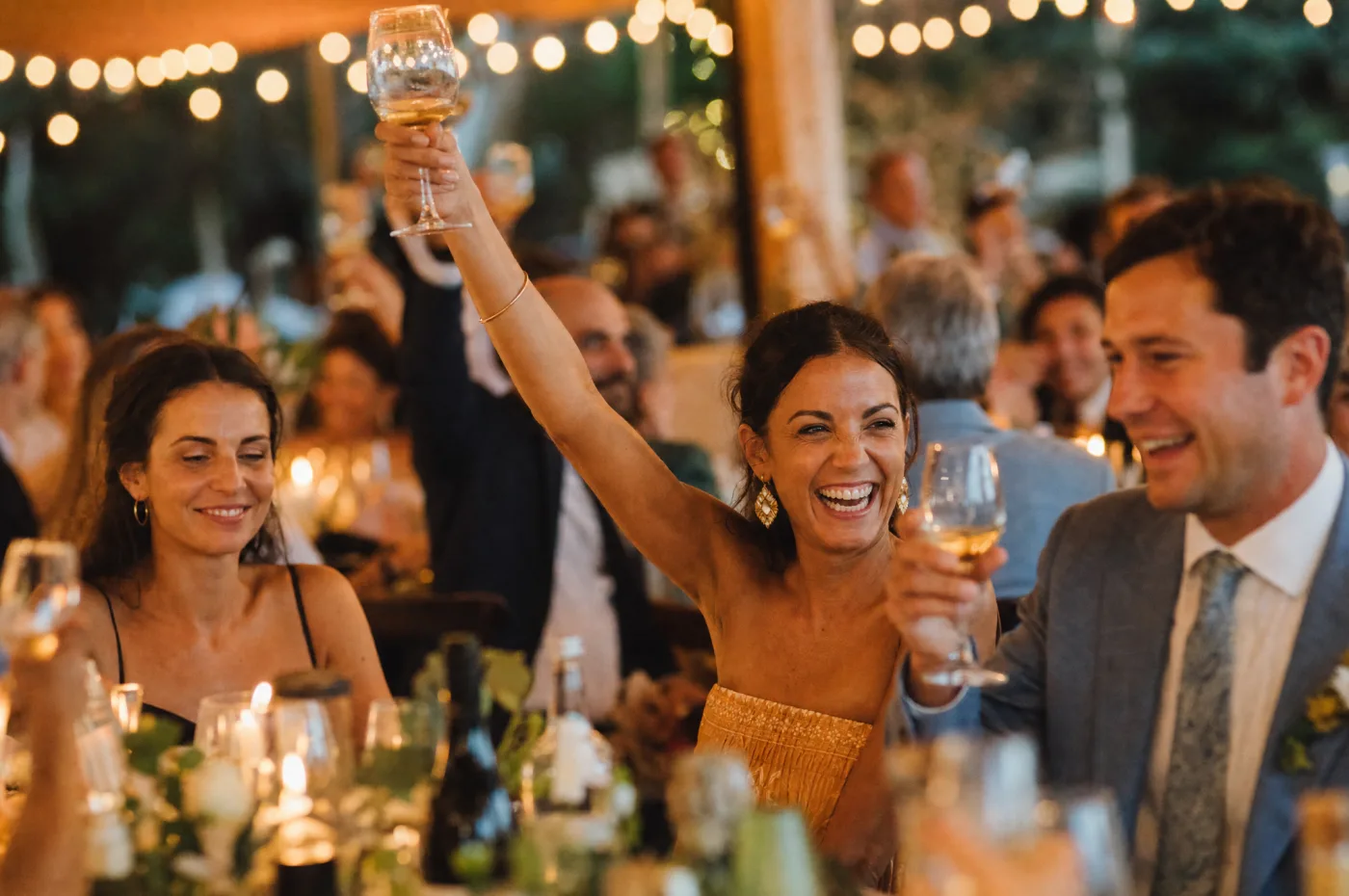 Guests laughing and raising a toast, string lights softly blurred behind them